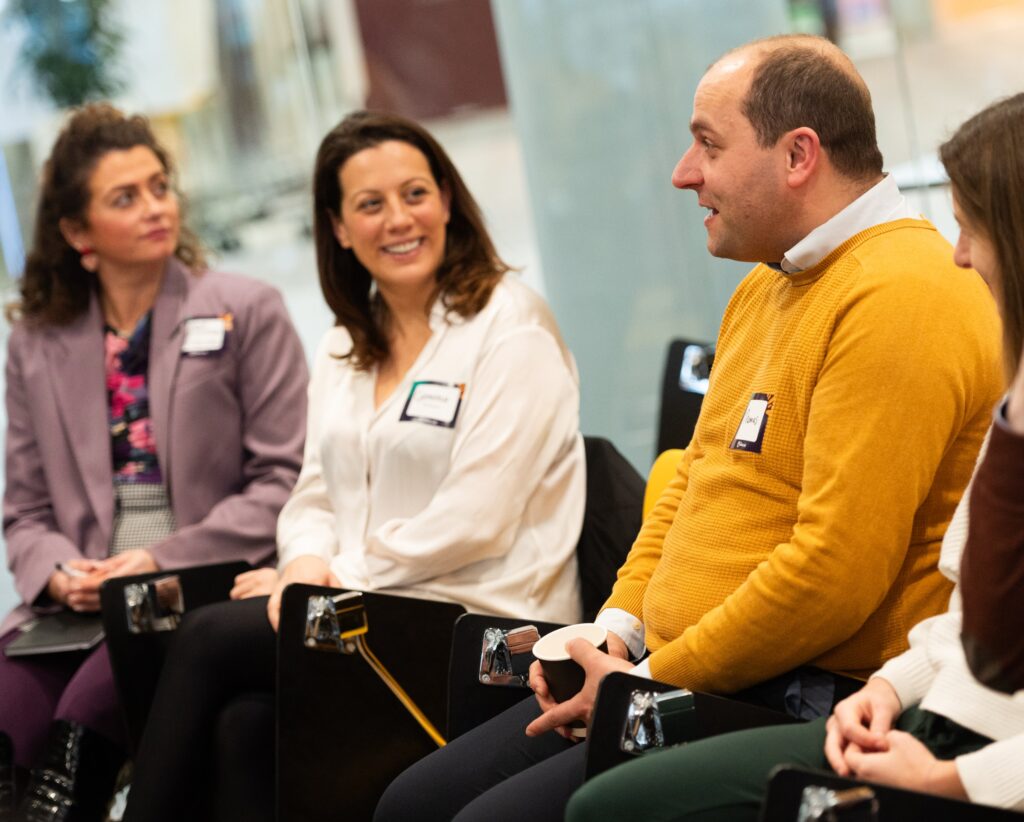 Two women smiling at a man in a yellow sweater who is talking. They are all sitting in a professional development workshop