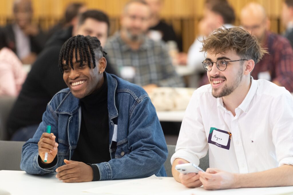 A young white male student with glasses and short brown and a young Black student sitting at a table and smiling brightly off to the left of the camera.