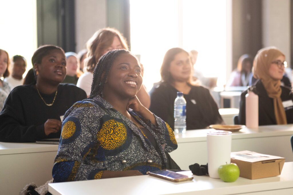 Woman smiling seated in the foreground of lecture theatre, rows of people in the background out of focus.